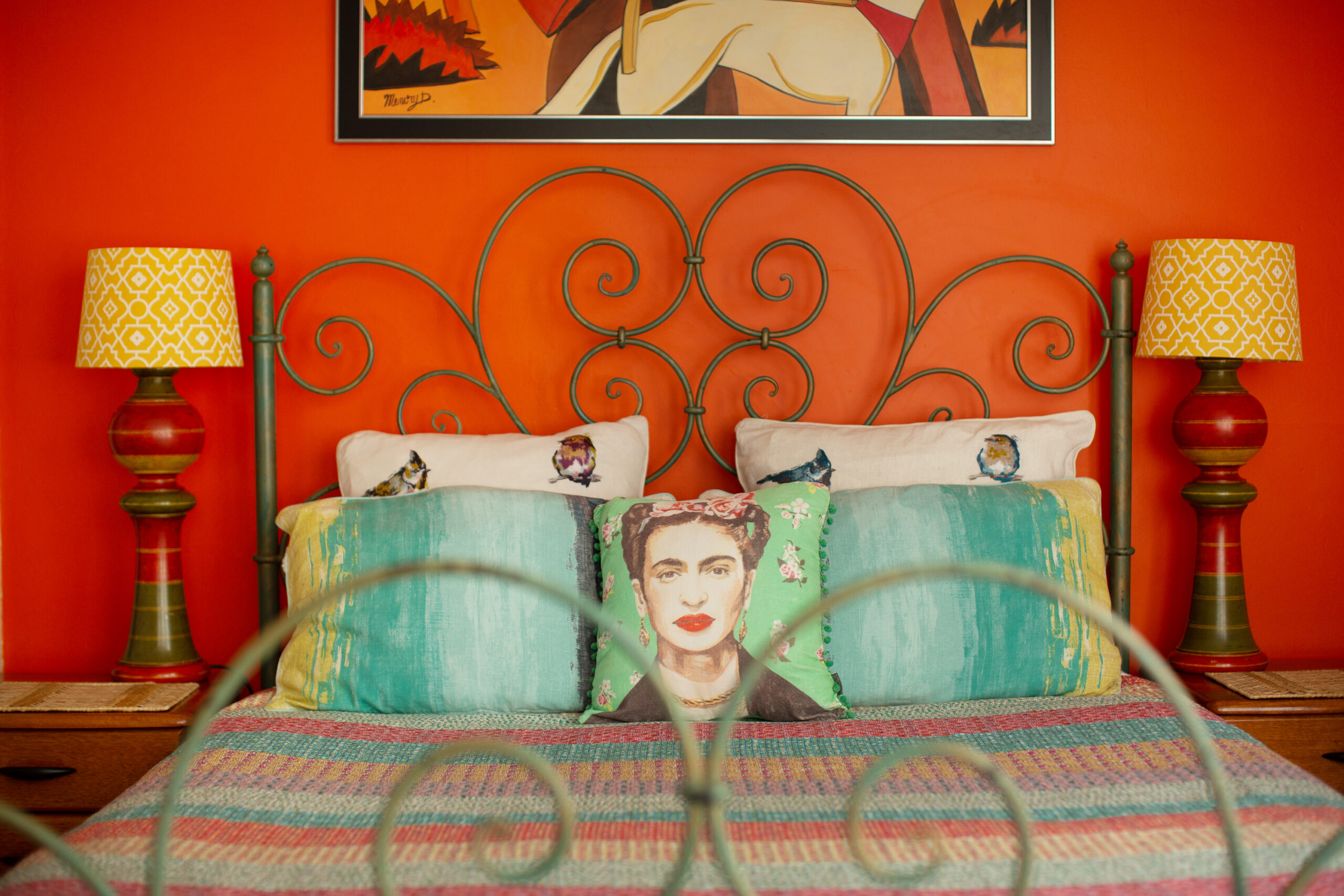 Photo of a bed against an orange wall. The pillow in the middle has a picture of Frida Kahlo on it.